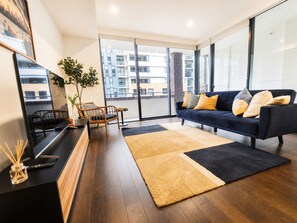 A modern living room with a dark blue sofa, a coffee table, and a large window with a city view. - Modern 2BDR Near Airport and Train Station (Mascot)