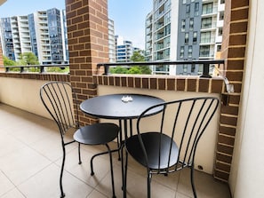 Ein Balkon mit rundem Tisch und zwei Stühlen, eine Ziegelwand und Blick auf hohe Gebäude.