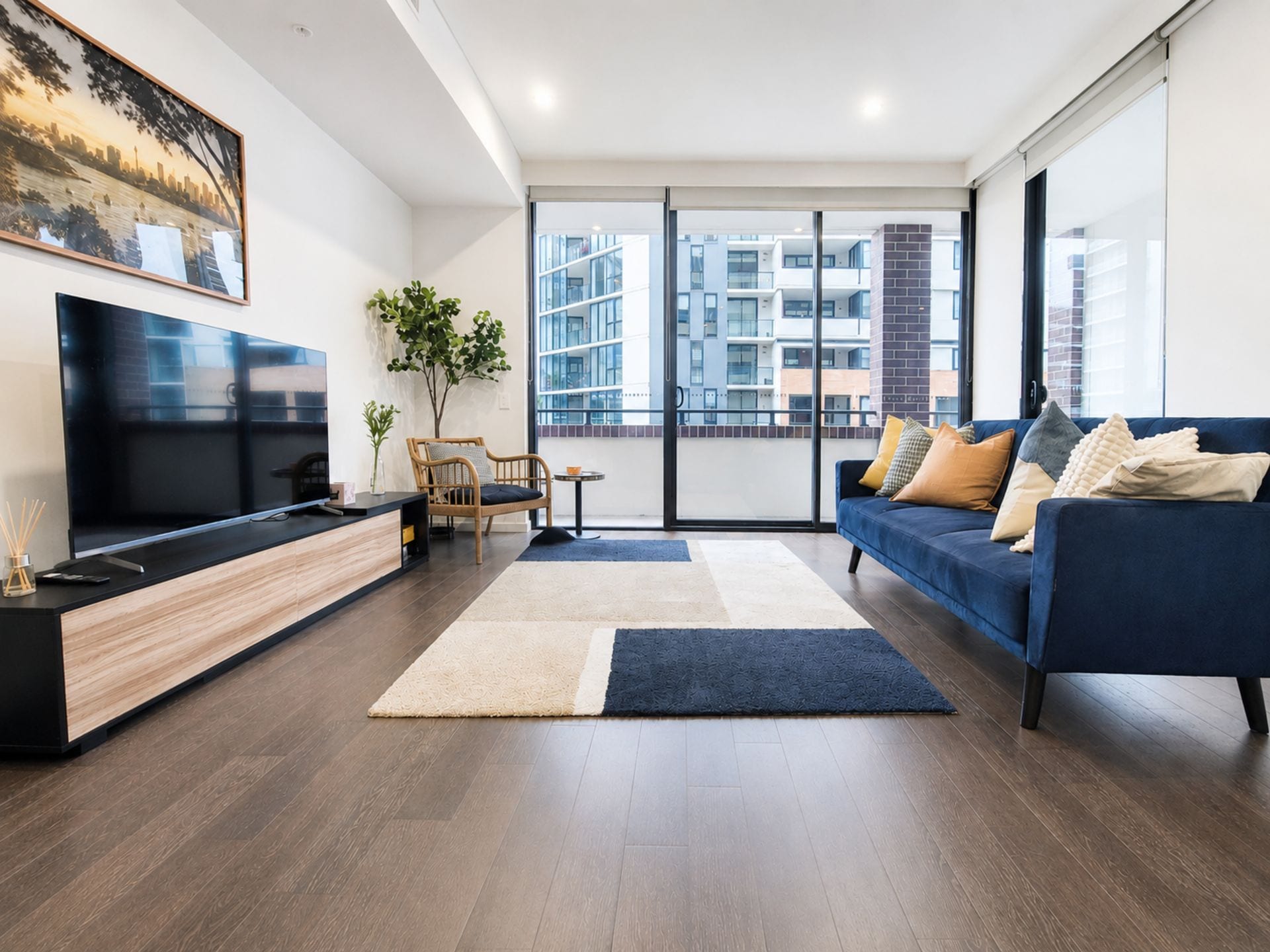 A modern living room with a flat-screen TV, a blue sofa with cushions, a wooden coffee table, and a large window with a city view.