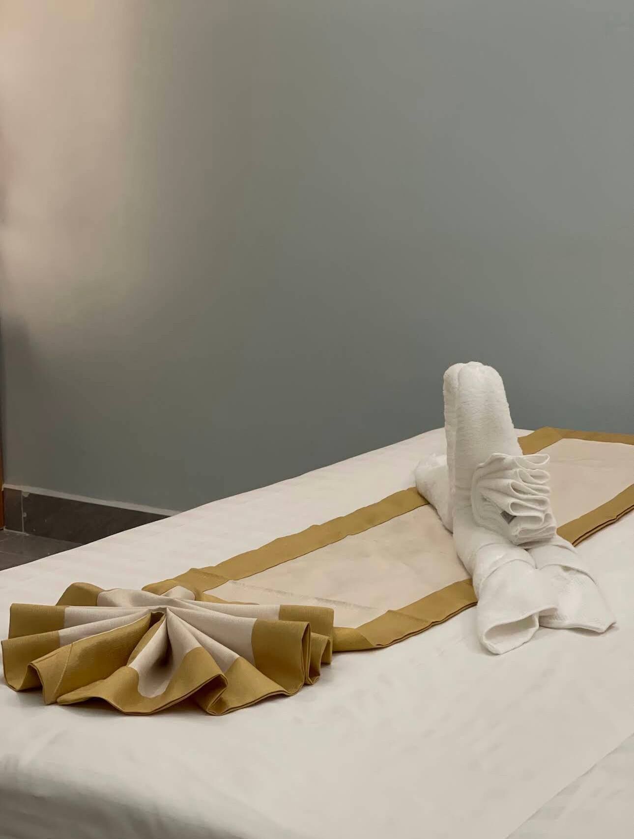A neatly made bed with a white and gold striped bedspread, a folded white towel, and a folded brown towel.