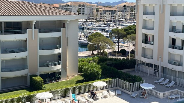 Un hotel de varios pisos con balcones, un puerto deportivo con barcos y una vista despejada de montañas al fondo.