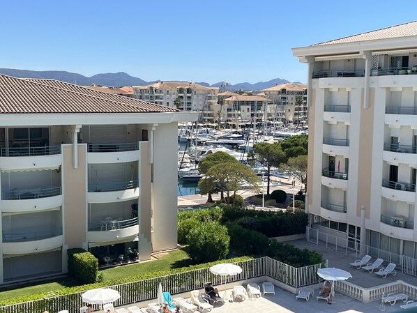 A multi-story hotel with balconies, a marina with boats, and a clear view of mountains in the background. - OPEN Residence, 1-bedroom apartment (41m²), sleeps 5, close to port, beaches, swimming pool, private parking (Fréjus)