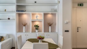 Moderni hotellihuone, jossa on ruokailutila, valkoinen sohva, hedelmäkulho pöydällä ja ruukkukasvi.