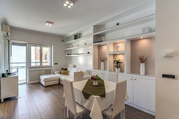 A modern living room with a dining area, white furniture, built-in shelves, and a view through large windows. - Olimpia, a luxury apartment in the heart of Monteverde in Rome (Roma)