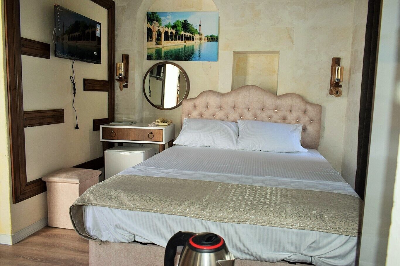A bedroom with a tufted headboard, a bed with patterned bedding, a bedside table with a lamp, a television mounted on the wall, and a framed picture above the bed.
