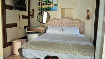 A bedroom with a tufted headboard, a bed with patterned bedding, a bedside table with a lamp, a television mounted on the wall, and a framed picture above the bed.