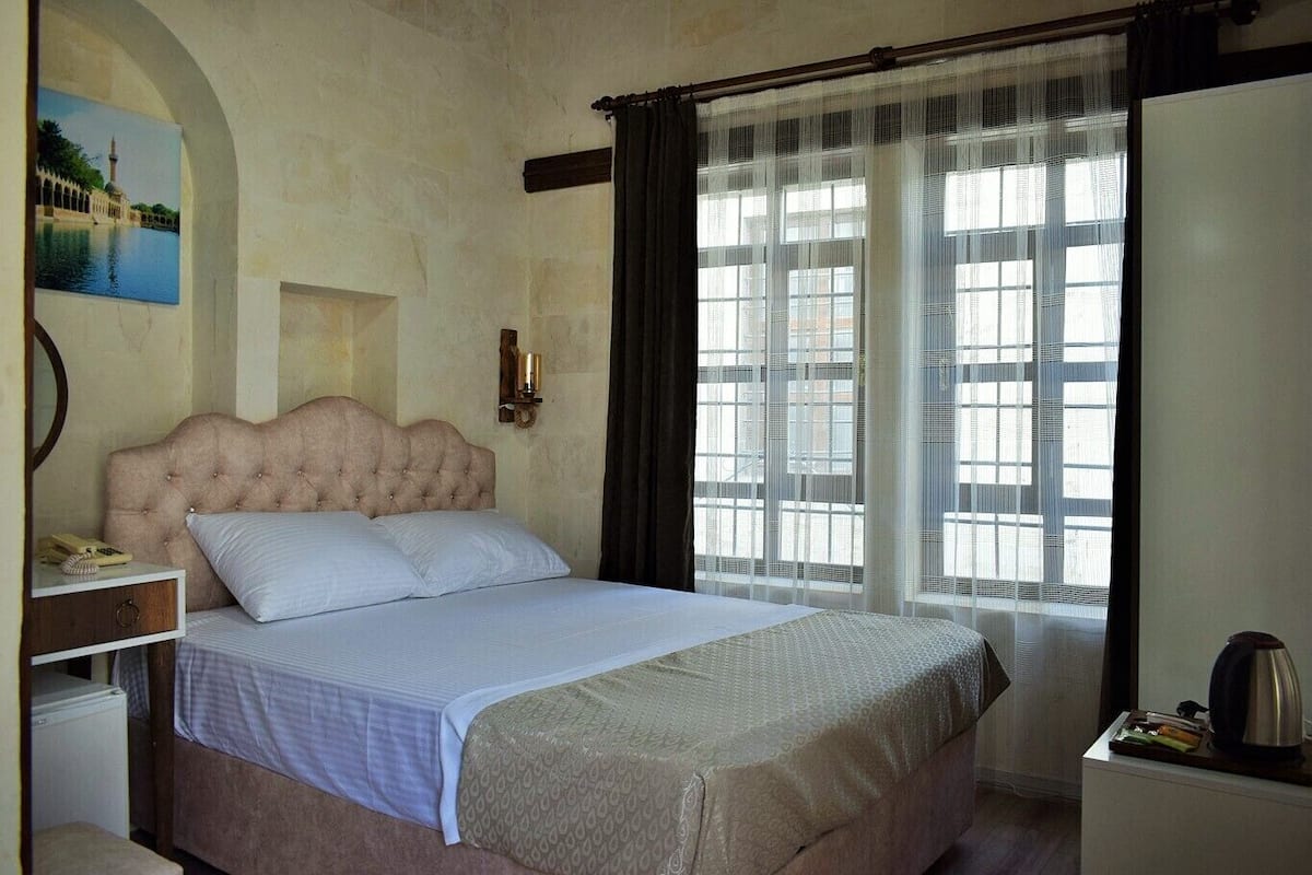 A bedroom with a large bed, a nightstand, a lamp, a window with curtains, and a painting on the wall.