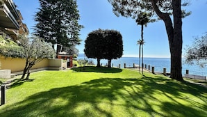 A well-maintained lawn with a large, neatly trimmed tree, a building with balconies, and a clear view of the sea. - Villa delle Rose Apartment 66 - Private Access to the Lake (Gardone Riviera)