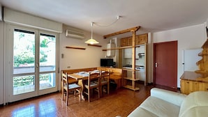 A room with a wooden dining table, chairs, a sofa, a television, and a kitchen area with wooden cabinets. - Villa delle Rose Apartment 66 - Private Access to the Lake (Gardone Riviera)
