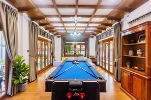Een pooltafel in een ruimte met houten balken, grote ramen en een kroonluchter.