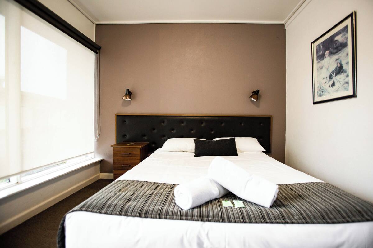 Ein Hotelzimmer mit einem großen Bett, einem Nachttisch, einem Fenster und einem gerahmten Bild an der Wand.