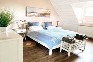 A bedroom with a sloped ceiling, a bed with a blue blanket, two bedside tables with lamps, a wall-mounted painting, and a potted plant. - Holiday home with 3 bedrooms (Wangerland\/Horumersiel\/Schillig)