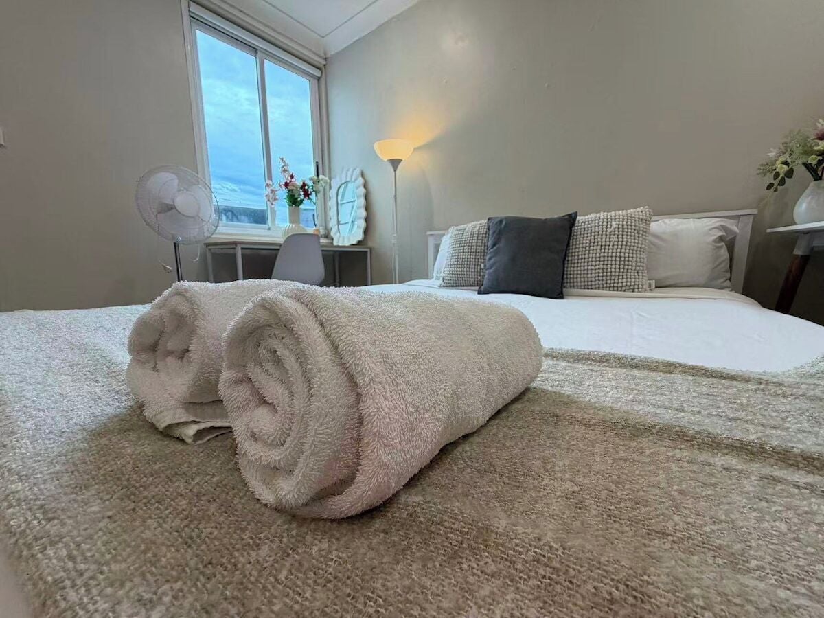 A neatly made bed with a rolled white towel in a room with a large window, a desk, and a lamp.