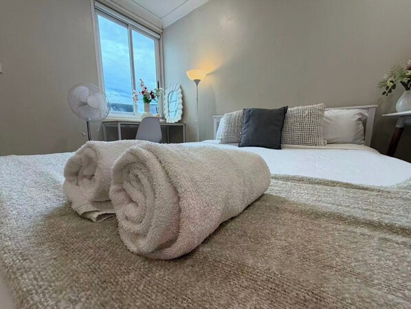 Ein ordentlich bezogenes Bett mit einem aufgerollten weißen Handtuch in einem Zimmer mit großem Fenster, Schreibtisch und Lampe.
