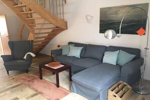 Een woonkamer met een grijze bank, een houten stoel, een klein tafeltje, een trap, een wandlamp en een schilderij.