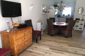 Een eetgedeelte met een houten tafel en rieten stoelen, een televisie aan de muur en een keuken op de achtergrond.