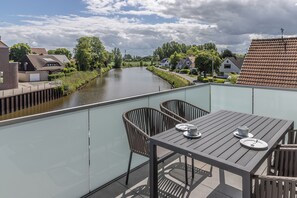 A balcony with a table and chairs, overlooking a canal with houses and greenery.