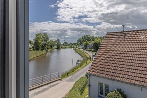Une rivière bordée d’une promenade dallée, des maisons aux toits recouverts de tuiles rouges et un ciel dégagé.