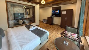Habitación de hotel con una cama grande, ducha con paredes de vidrio, televisión, un escritorio pequeño y una mesa de comedor.