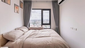 Ein Schlafzimmer mit einem Bett, einem Fenster mit Vorhängen, einer Klimaanlage und Blick auf die Stadt.
