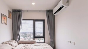 Ein Schlafzimmer mit einem Bett, einem Fenster mit Vorhängen, einer Klimaanlage und Blick auf die Stadt.