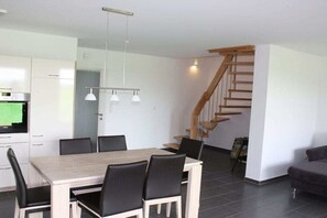 Eine moderne Küche mit weißen Schränken, einem Esstisch mit Stühlen, Pendelleuchten und einer Treppe.