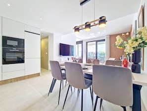 Eine moderne Küche mit integrierten Geräten, ein Essbereich mit Stühlen und Tisch sowie ein großer Flachbildfernseher an der Wand.
