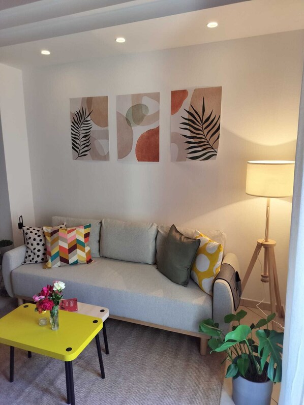 A living room with a sofa, colorful cushions, a small yellow table with flowers, and a potted plant.