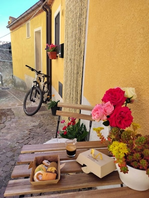 Un banc en bois, une boîte de pâtisseries, une tasse de café et un vélo appuyé contre un immeuble jaune.