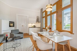 Eine moderne Küche mit weißen Schränken, ein Essbereich mit weißem Tisch und Stühlen sowie ein großes Fenster mit Blick auf Gebäude.