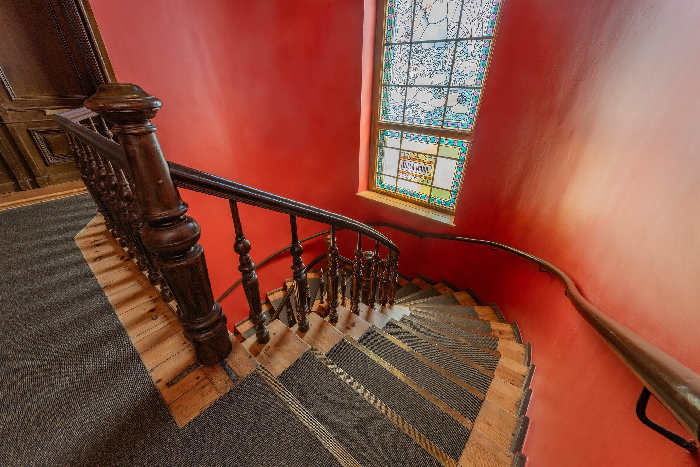 Un escalier en bois avec une rampe, des marches recouvertes de moquette et un vitrail portant l’inscription « VILLA MARIE ».