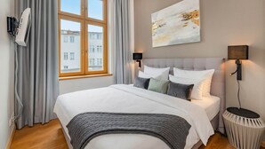 Quarto com uma cama grande, janela com cortinas, ar condicionado fixado na parede, mesa lateral redonda e um quadro na parede.