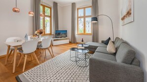 Uma sala de estar moderna com um sofá cinzento, uma mesa de jantar com cadeiras, uma televisão e uma janela grande com cortinas.