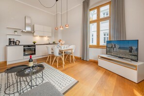 Eine moderne Küche mit weißen Schränken, ein Essbereich mit Tisch und Stühlen, ein Fernseher auf einem Schrank und ein Fenster mit Vorhängen.