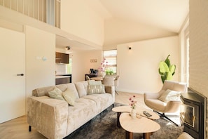 Ein Wohnzimmer mit Sofa, Sessel, Couchtisch, Kamin und einer Küche im Hintergrund.