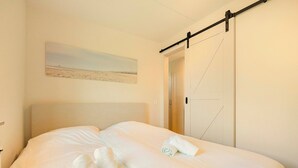 Un lit bien fait, avec du linge de lit blanc et des serviettes pliées, un tableau représentant une scène de plage accroché au mur, et une porte coulissante sur le côté droit.