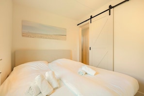 Ein ordentlich bezogenes Bett mit weißen Leintüchern und aufgerollten Handtüchern, ein Strandbild an der Wand und eine Schiebetür rechts.