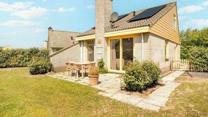Une maison avec une façade en briques, une cheminée, un panneau solaire, un patio avec une table et des chaises, et un jardin.