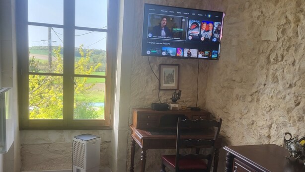 Et værelse med et TV ophængt på væggen, et træbord, en stol, en radiator og et vindue med udsigt til grøn vegetation.