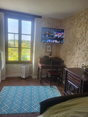Quarto com televisão na parede, uma escrivaninha de madeira, uma cadeira, radiador e janela com vista para área verde.
