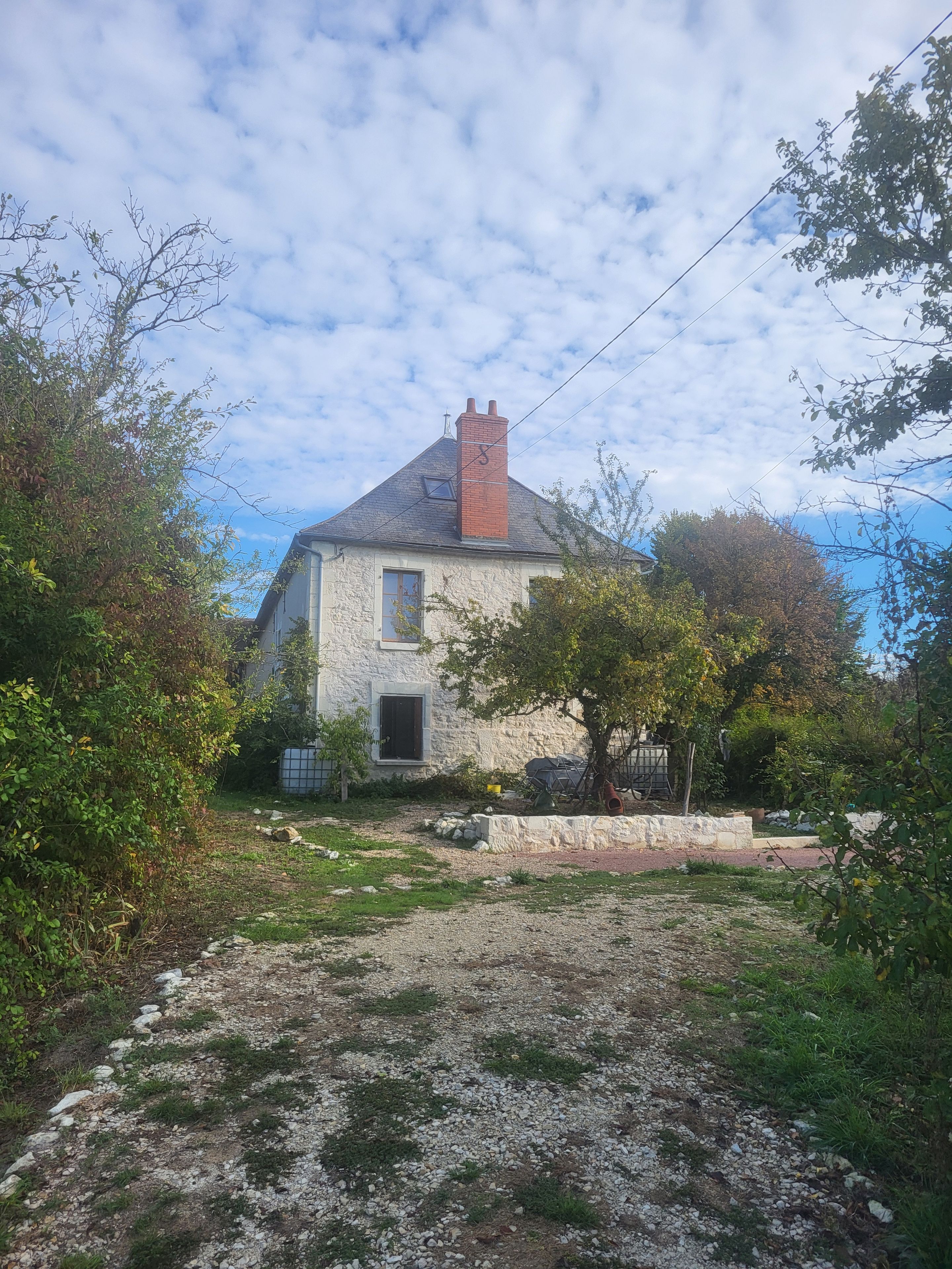 A stone house with a chimney, a garden with a gravel path, and a tree in the foreground.