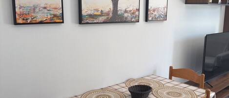 A dining area with a table set for four, a television on a stand, and three abstract paintings on the wall.