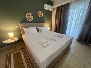 Ein Hotelzimmer mit einem großen Bett, zwei Wand-Klimaanlagen, einer Nachttischlampe und Blick ins Freie durch Vorhänge.