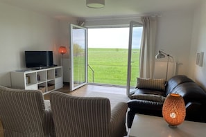 Ein Wohnzimmer mit Fernseher, Sofa, Sesseln, einer Lampe und Blick durch eine Glastür.