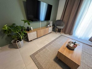 Ein modernes Wohnzimmer mit Flachbildfernseher, Couchtisch aus Holz, grauem Sessel, Blumentopf und einer Ablage mit Dekoration.