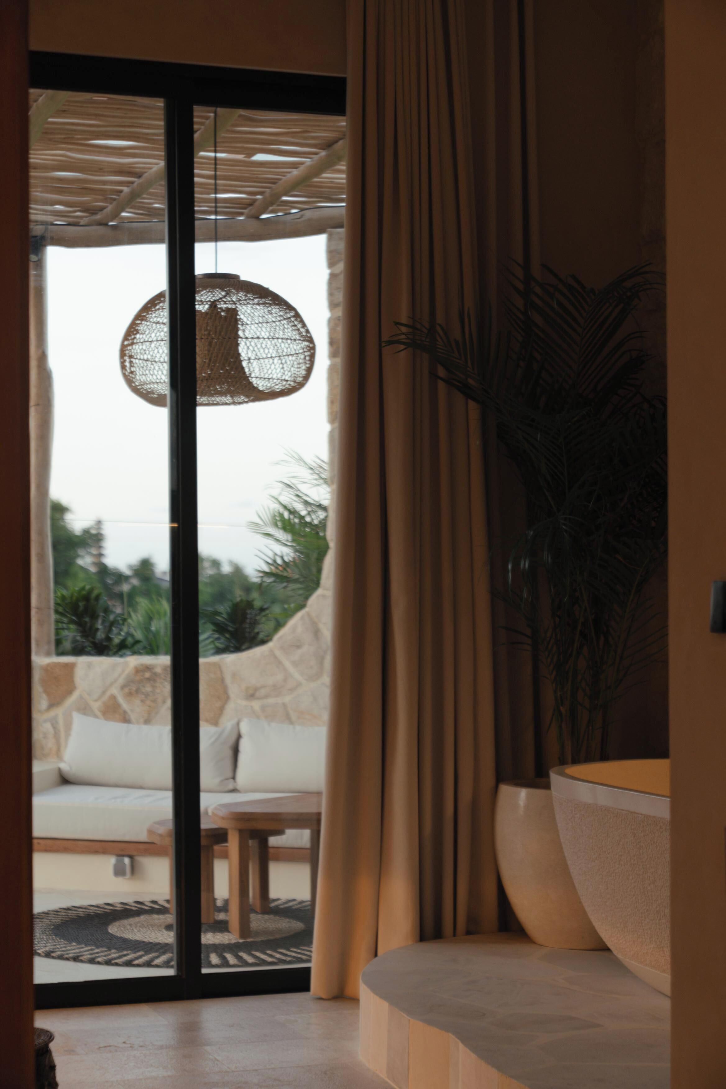 A room with a sliding glass door leading to an outdoor area, a woven light fixture, a large plant, and a modern sofa.