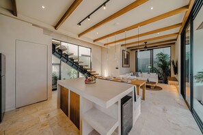 A modern kitchen with a central island, a dining area with a sofa and table, and a staircase leading to an upper level. - Maison Kala (Cemagi)
