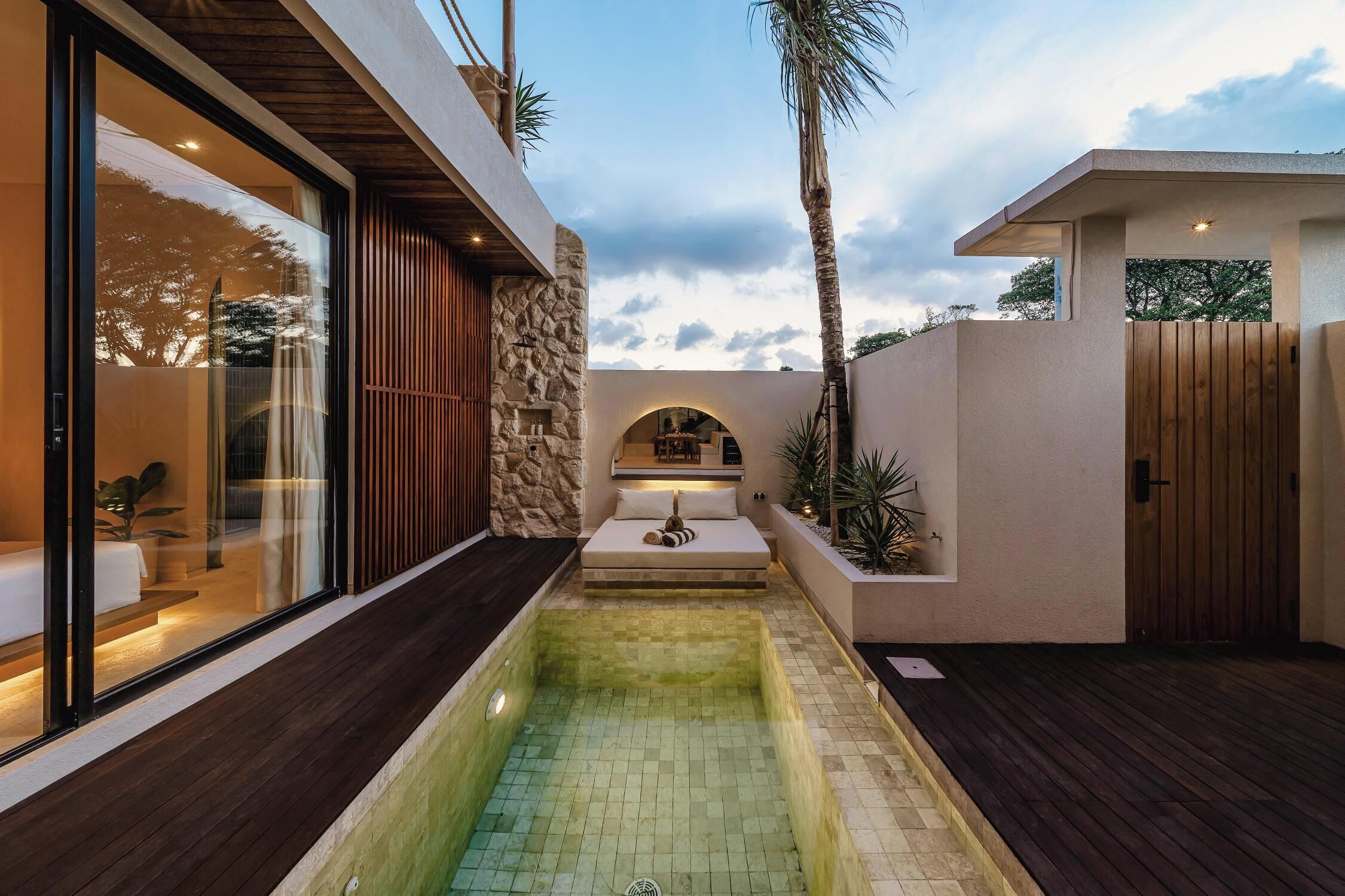 A modern outdoor pool area with a stone wall, wooden deck, and a glass-enclosed room.