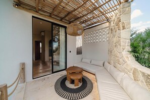 Ein modernes Zimmer mit einer Steinwand, einer gewebten Decke, einer Glastür, die auf einen Balkon führt, einem runden Holztisch und einem weißen Sofa.