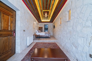 A narrow hallway with a wooden door, a bed with a view, stone walls, and a patterned rug.